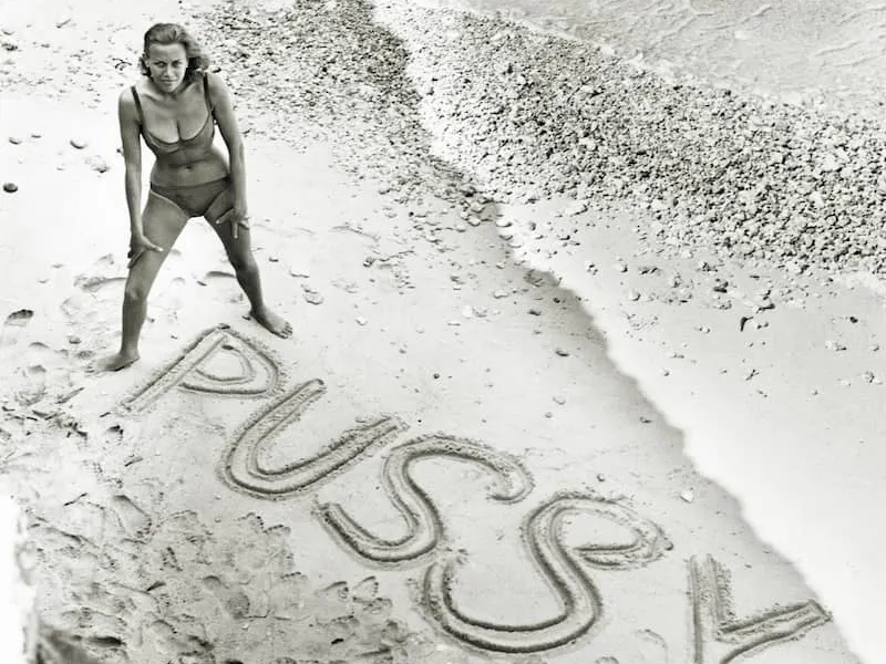 Honor Blackman standing on a beach with Pussy Galore written in the sand, Goldfinger publicity photo