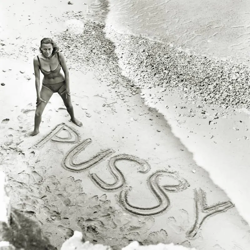 Honor Blackman standing on a beach with Pussy Galore written in the sand, Goldfinger publicity photo